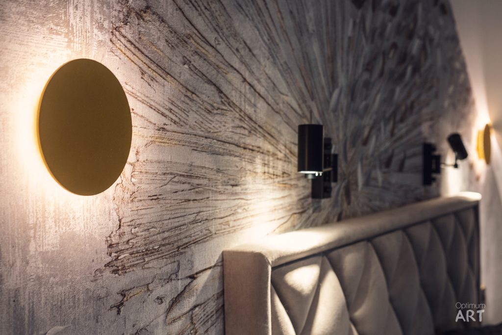 Nowoczesne wnętrze schodów z charakterystyczną okrągłą lampą LED na teksturowanej ścianie i eleganckimi balustradami. Ciepłe oświetlenie podkreśla fakturę ściany i tworzy nastrojową atmosferę w przestrzeni.