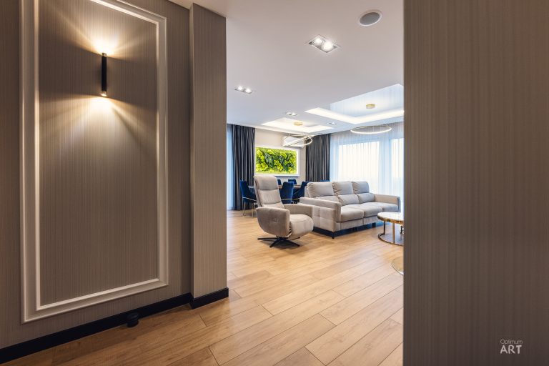 Eleganckie wnętrze mieszkania z jasnym drewnianym parkietem, gdzie z przedpokoju widać salon z szarymi meblami wypoczynkowymi i niebieskimi zasłonami. Na ścianie w przedpokoju wisi nowoczesna lampa, a całość utrzymana jest w stonowanej kolorystyce beży i szarości.