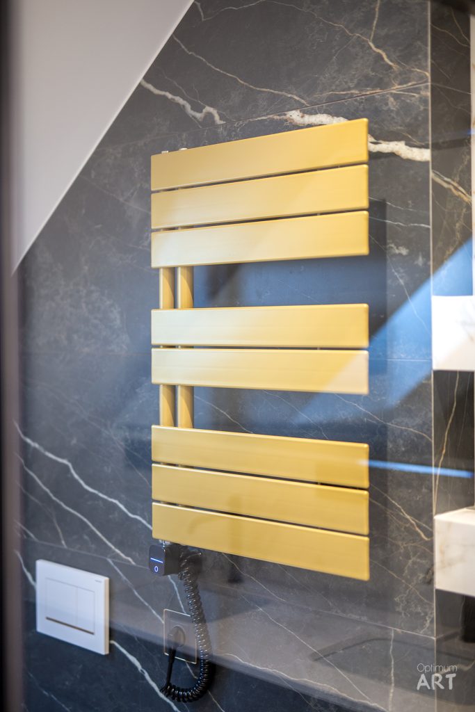 Nowoczesny grzejnik łazienkowy w kolorze złotym zamontowany na ścianie z ciemnego marmuru z białymi żyłkami. Grzejnik ma poziome płaskie listwy połączone pionowymi rurkami, a obok widoczny jest włącznik światła.