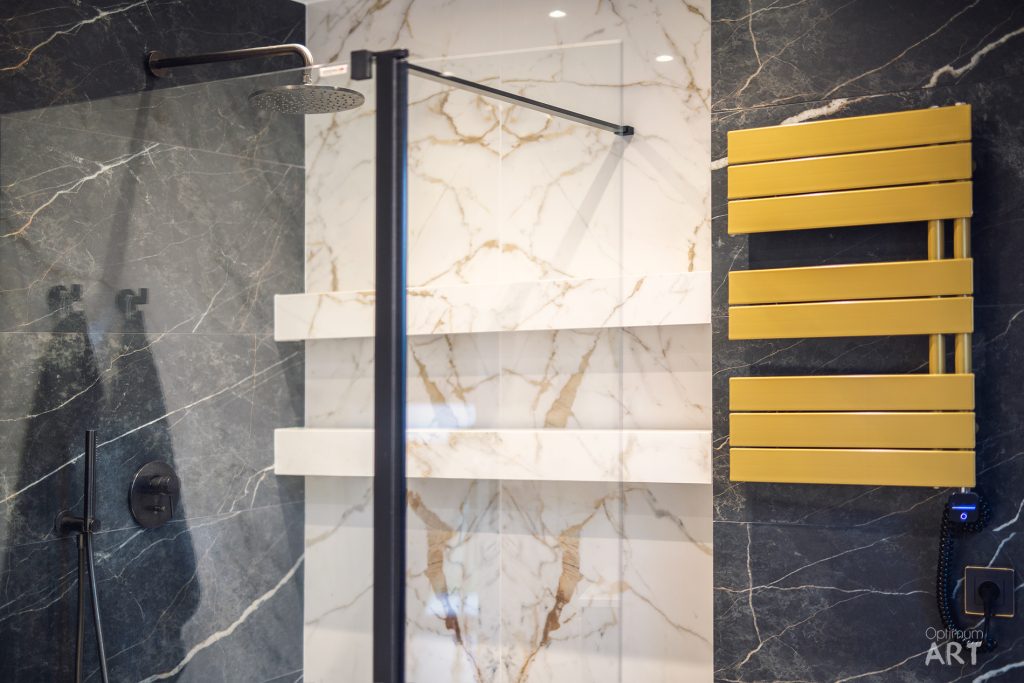 Nowoczesna łazienka z marmurowymi ścianami, w której znajduje się kabina prysznicowa z czarną armaturą, przezroczystą szybą oraz żółty grzejnik łazienkowy zamontowany na ścianie.