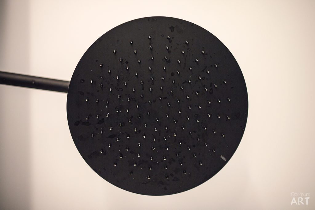 Czarna okrągła deszczownica prysznicowa z licznymi małymi otworami, zamontowana na ciemnej rurze przy jasnej ścianie. W prawym dolnym rogu widoczny jest napis "Optimum ART".