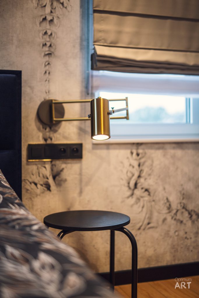 Nowoczesne wnętrze sypialni z złotą lampą ścienną na wysięgniku, czarnym okrągłym stolikiem i łóżkiem z wzorzystą pościelą. W tle widoczna tapeta z delikatnym wzorem roślinnym i okno z roletami.