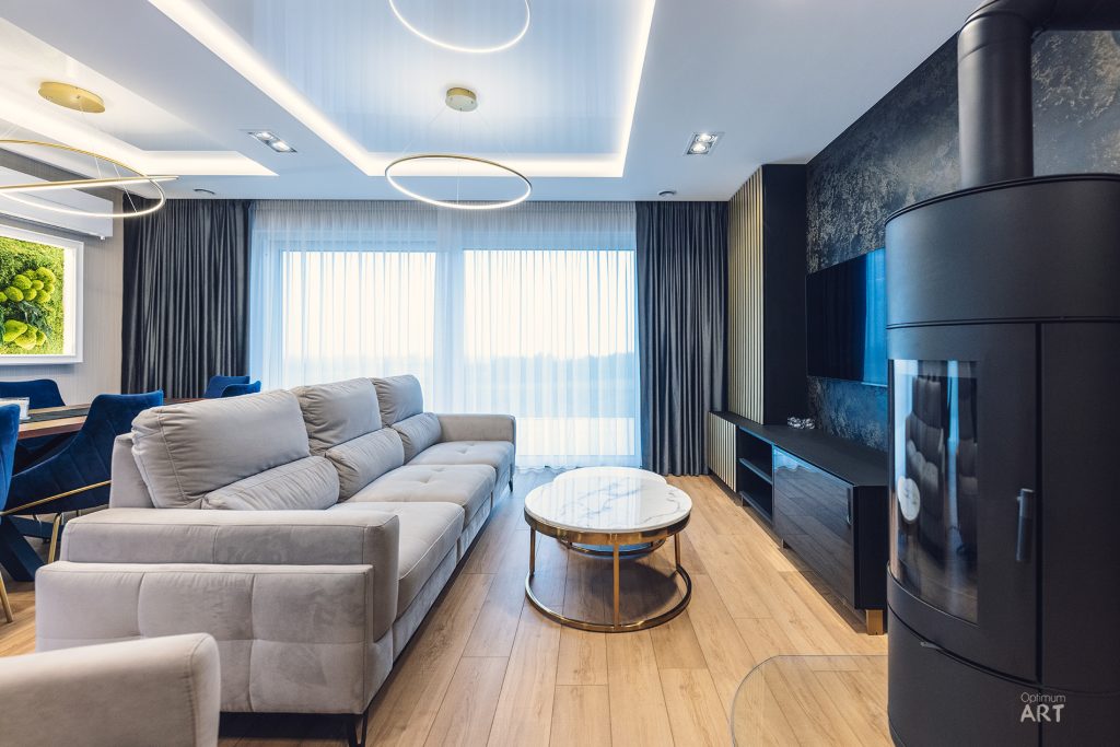 Nowoczesny salon z szarą sofą narożną, okrągłym stolikiem kawowym i czarnym kominkiem. Pomieszczenie ma drewnianą podłogę, duże okna z zasłonami oraz nowoczesne oświetlenie sufitowe.