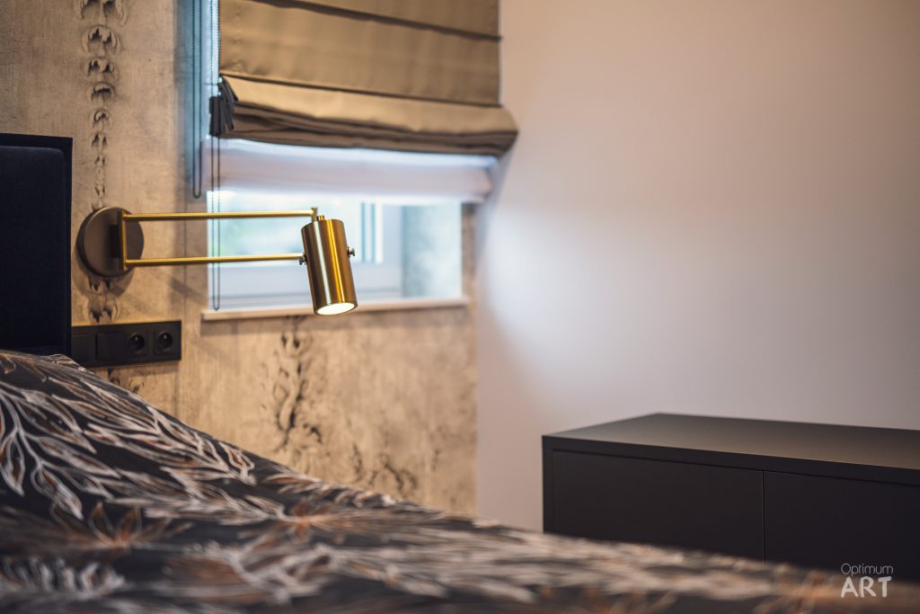 Sypialnia z łóżkiem przykrytym wzorzystą pościelą, przy którym na ścianie zamontowana jest złota lampka do czytania na wysięgniku. Obok łóżka znajduje się ciemna szafka nocna, a w tle widać okno z roletą i fragment telewizora.