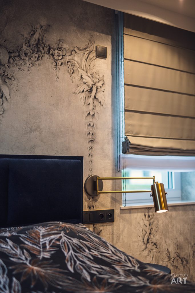 Sypialnia z łóżkiem przykrytym wzorzystą pościelą, złotą lampą ścienną i dekoracyjną ścianą z reliefowymi ornamentami roślinnymi. Obok łóżka znajduje się okno z beżowymi żaluzjami poziomymi.