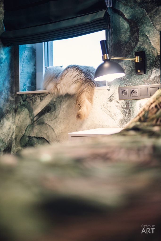 Puszysta koty ryży kot leży na białym parapecie przy oknie w łazience z kamienną ścianą. Nad kotem wisi czarna lampa ścienna, a na ścianie widoczne są gniazdka elektryczne.