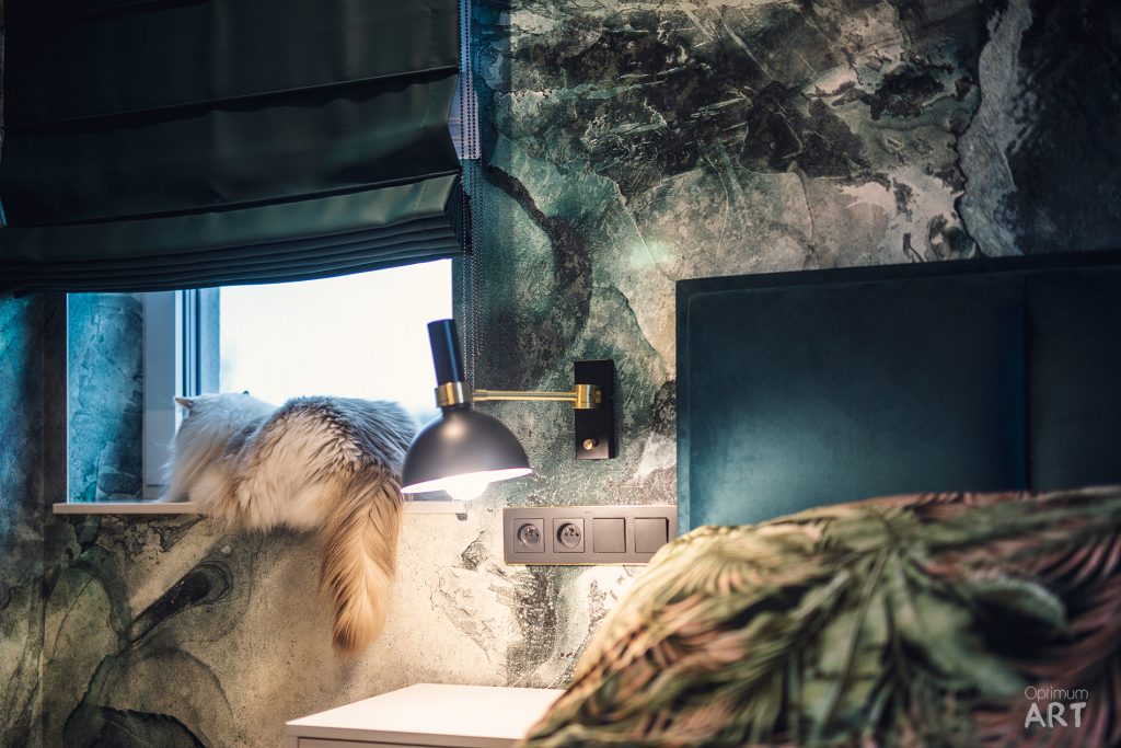 Nowoczesna sypialnia z efektowną ścianą pokrytą naturalnym kamieniem lub marmurem w odcieniach zieleni i beżu. Nad łóżkiem wisi czarna lampa wisząca, a obok okna widoczne są dekoracyjne trawy i rośliny, które dopełniają naturalnego charakteru wnętrza.