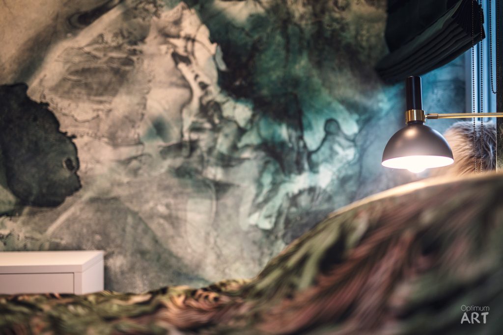 Nowoczesne wnętrze z charakterystyczną ścianą pokrytą marmurowymi wzorami w odcieniach zieleni i beżu. W prawej części widoczna elegancka lampa wisząca z mosiężnymi elementami oraz fragment drewnianego blatu.