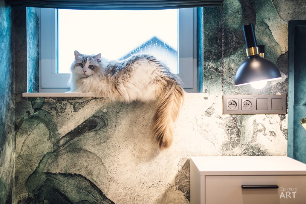 Puszysty kot o jasnym futerku z ciemnymi pasami siedzi na parapecie przy oknie w nowoczesnym wnętrzu z kamienną ścianą. Obok kota widoczna jest stylowa lampa wisząca i gniazdka elektryczne wmontowane w ścianę.