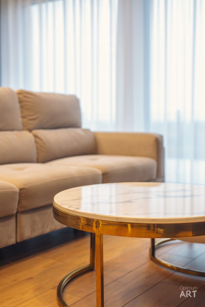 Beżowa sofa narożna stoi przy oknie z białymi zasłonami, a przed nią znajduje się okrągły stolik kawowy ze złotą metalową ramą i marmurowym blatem. Całość umieszczona jest na drewnianej podłodze w jasnym, nowoczesnym salonie.