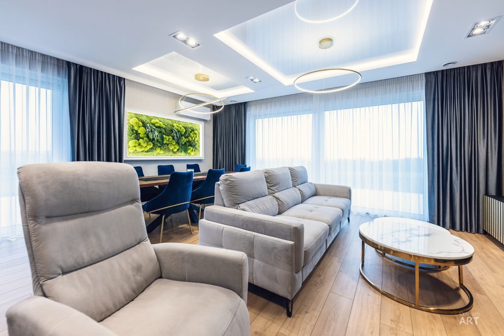 Nowoczesny salon z szarą sofą narożną i fotelem, okrągłym stolikiem kawowym oraz stołem jadalnym z niebieskimi krzesłami w tle. Na ścianie wisi duży obraz z zielonymi roślinami, a pomieszczenie ma stylowy sufit z oświetleniem LED i duże okna z ciemnoniebieskimi zasłonami.
