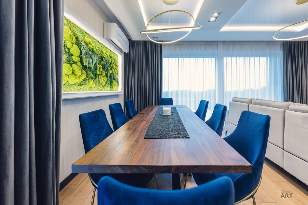 Nowoczesna jadalnia z długim drewnianym stołem otoczonym niebieskimi krzesłami, na ścianie wisi duży obraz z zieloną roślinnością. Po prawej stronie znajduje się jasna sofa, a pomieszczenie oświetlają nowoczesne lampy wiszące i naturalne światło z okna.