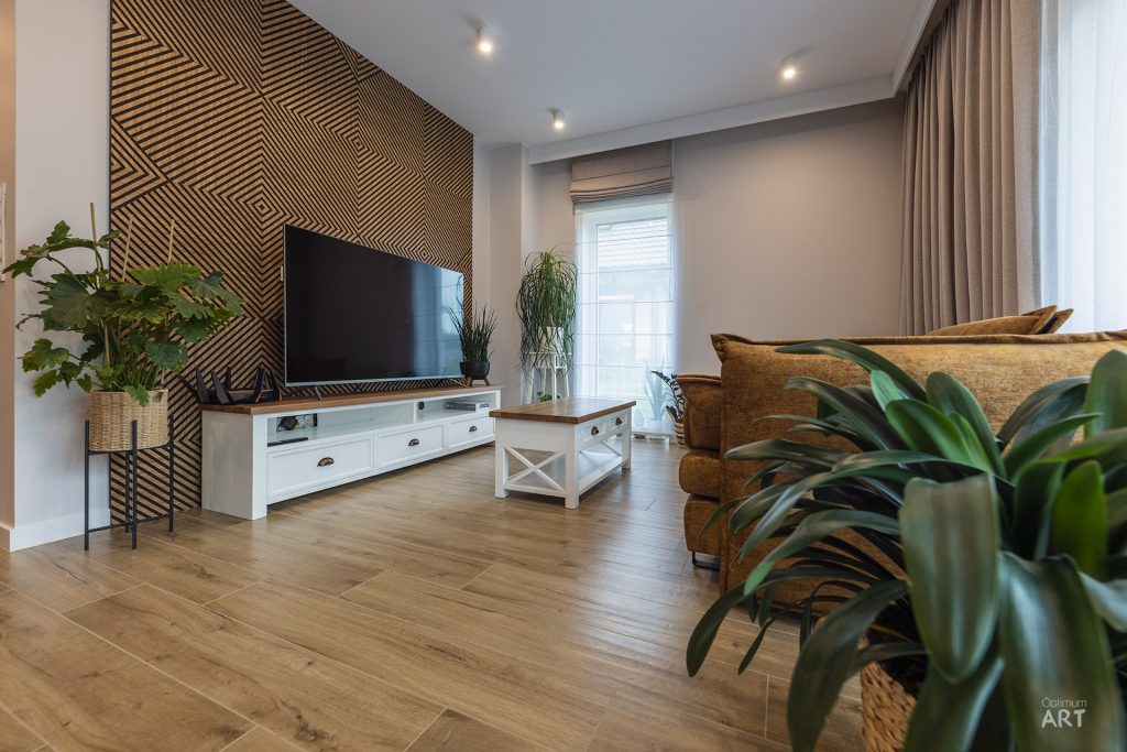 Nowoczesny salon z drewnianą podłogą, białą szafką pod telewizorem i charakterystyczną ścianą z geometrycznym wzorem z drewnianych listewek. W pomieszczeniu znajdują się rośliny doniczkowe, brązowa sofa oraz duże okno z zasłonami.