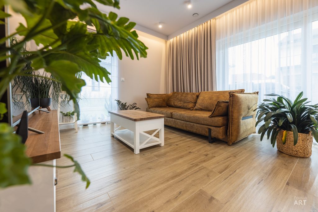 Jasny salon z brązową sofą i białym stolikiem kawowym, otoczony licznymi zielonymi roślinami doniczkowymi. Pomieszczenie ma drewnianą podłogę, beżowe zasłony w dużym oknie i nowoczesne oświetlenie.