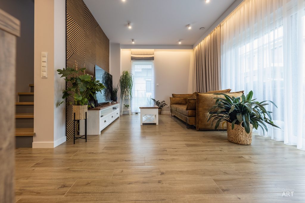 Nowoczesny salon z drewnianą podłogą, brązową sofą i białą szafką RTV, oświetlony naturalnym światłem z dużego okna z zasłonami. Na ścianie widoczne są drewniane panele dekoracyjne, a w pomieszczeniu ustawiono kilka dużych roślin doniczkowych.