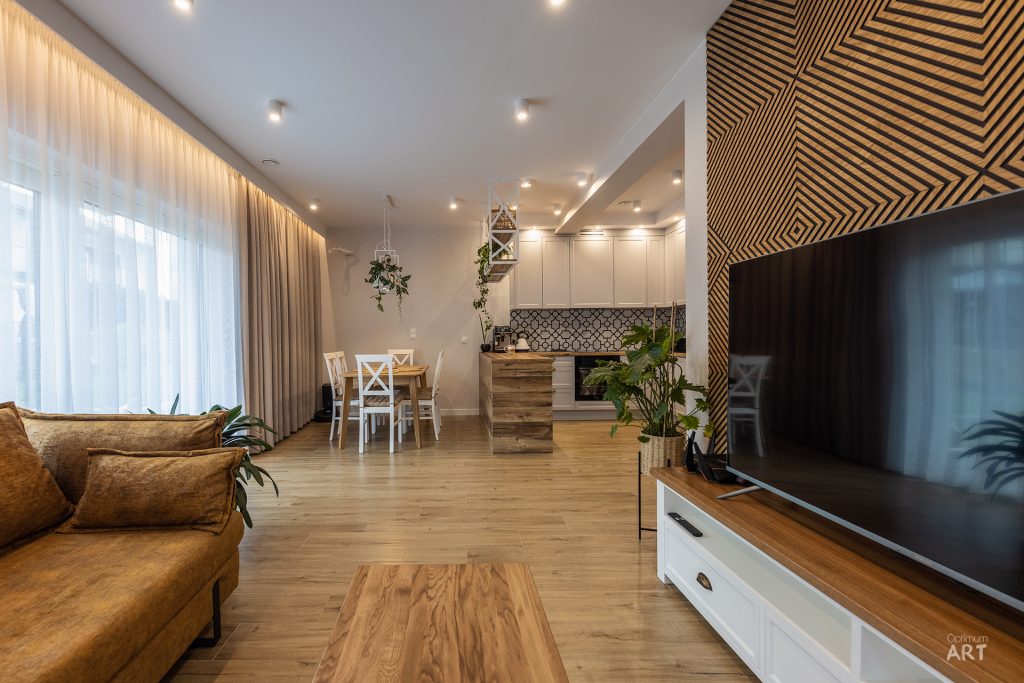 Nowoczesne wnętrze salonu z kuchnią w stylu skandynawskim, z brązową sofą, drewnianą podłogą i białymi meblami. Na ścianie za telewizorem znajduje się dekoracyjna drewniana boazeria w geometryczny wzór, a w tle widać jadalnię ze stołem i krzesłami.