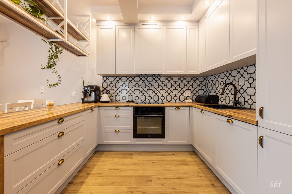 Nowoczesna kuchnia w kształcie litery U z białymi szafkami, drewnianymi blatami i geometrycznym wzorem na ścianie między blatem a szafkami górnymi. Na podłodze ułożone są jasne deski drewniane, a nad blatem wiszą otwarte półki z roślinami.