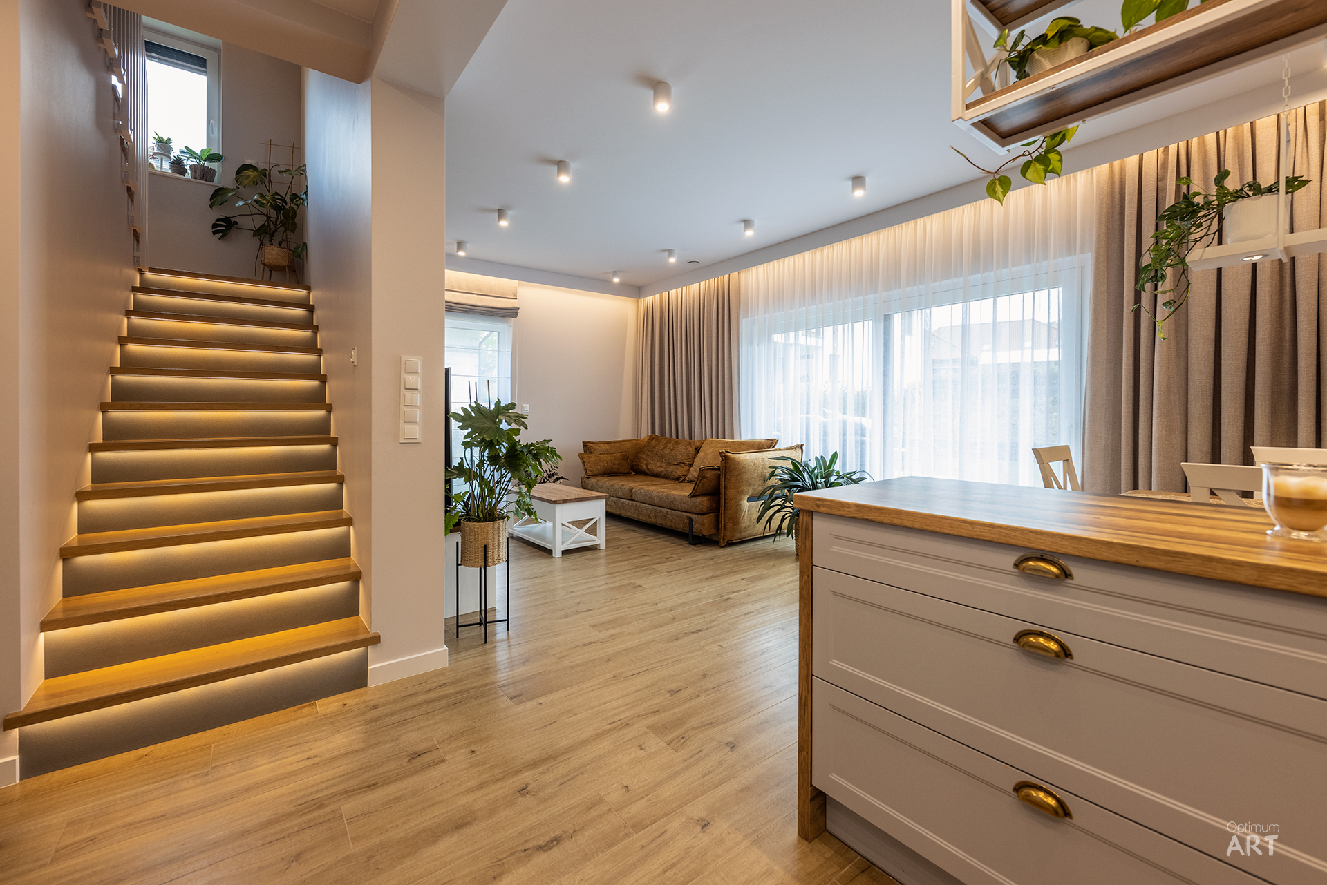Nowoczesne wnętrze salonu z drewnianymi schodami z podświetleniem LED po lewej stronie i beżową sofą przy oknie z zasłonami. Na pierwszym planie widać drewnianą komodę z szufladami, a całe pomieszczenie ozdobione jest zielonymi roślinami doniczkowymi.