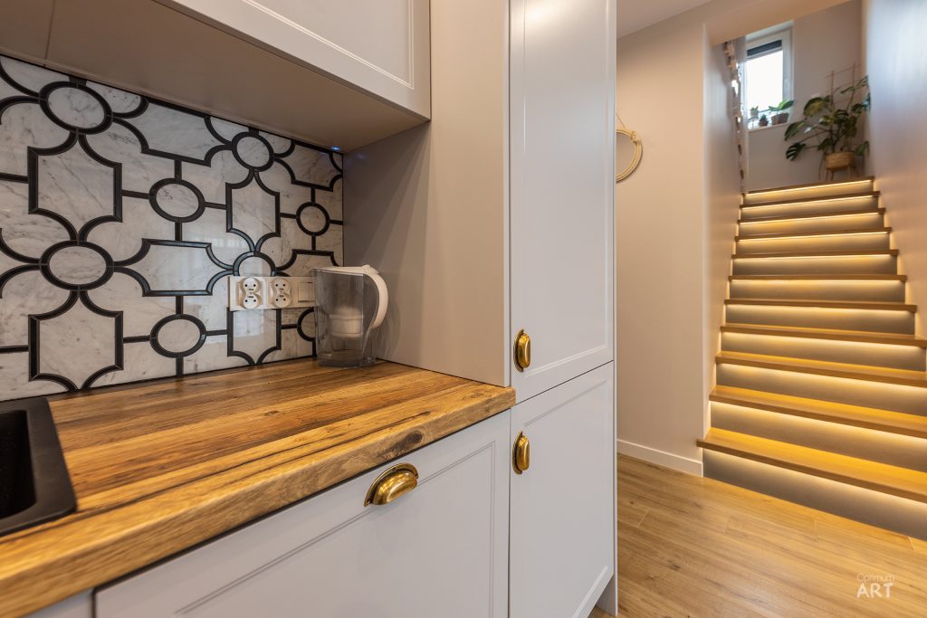 Nowoczesna kuchnia z białymi szafkami, drewnianym blatem i dekoracyjnymi płytkami na ścianie w geometryczne wzory. W tle widoczne są podświetlone schody z drewna prowadzące na górę.
