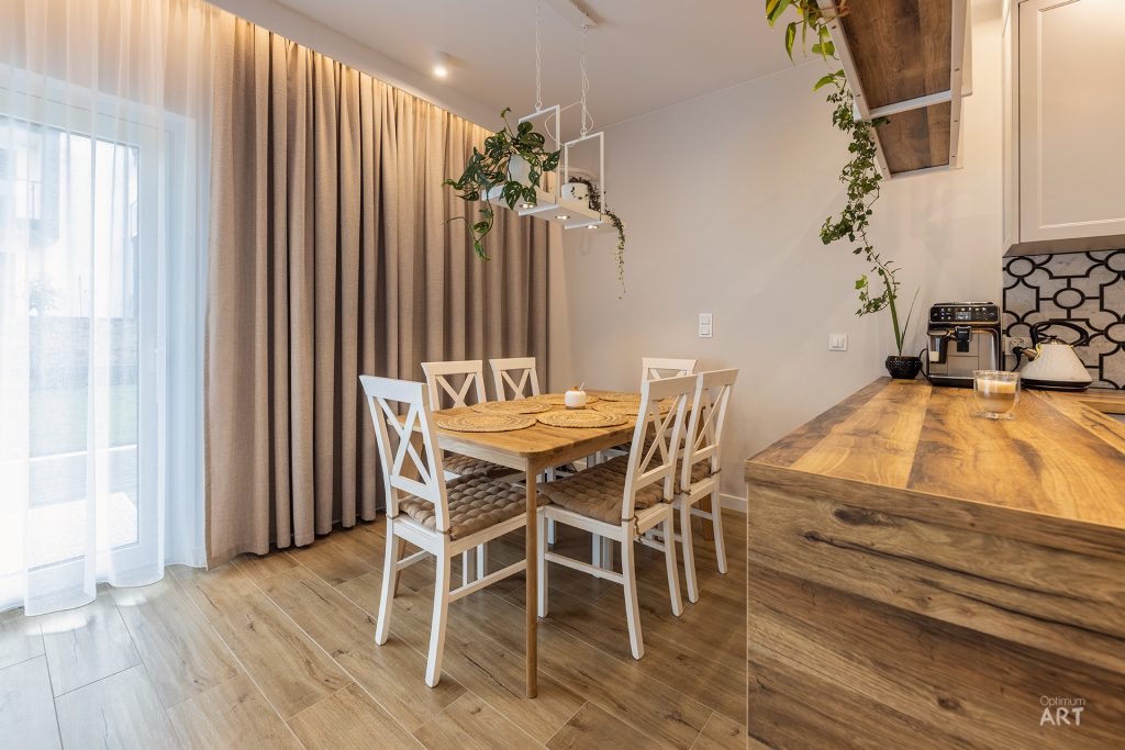 Jasna jadalnia z drewnianym stołem otoczonym białymi krzesłami o skrzyżowanych oparciach, połączona z kuchnią z drewnianym blatem. W tle beżowe zasłony przy oknie i wiszące rośliny doniczkowe.