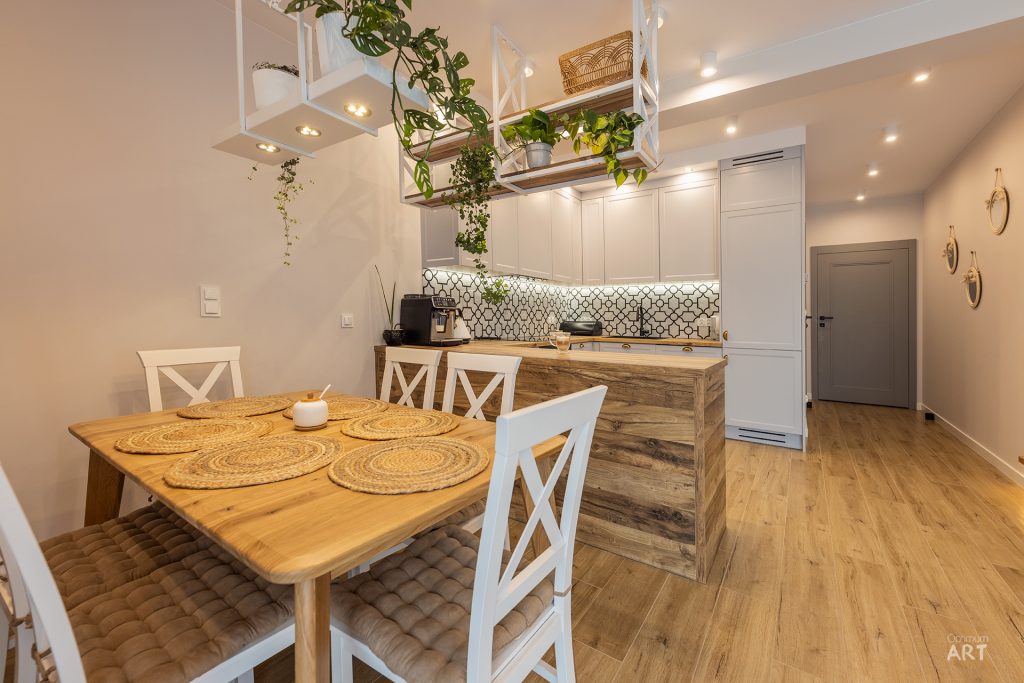 Nowoczesna kuchnia otwarta na jadalnię z drewnianym stołem i białymi krzesłami, wyposażona w białe szafki i drewniany blat. Nad wyspą kuchenną wiszą białe półki z roślinami doniczkowymi, a wnętrze ma ciepłe, beżowe ściany i drewnianą podłogę.