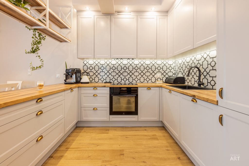 Nowoczesna kuchnia w kształcie litery U z białymi szafkami, drewnianymi blatami i złotymi uchwytami. Na ścianie znajduje się geometryczna mozaika w czarno-białych wzorach, a podłoga wykonana jest z jasnego drewna.