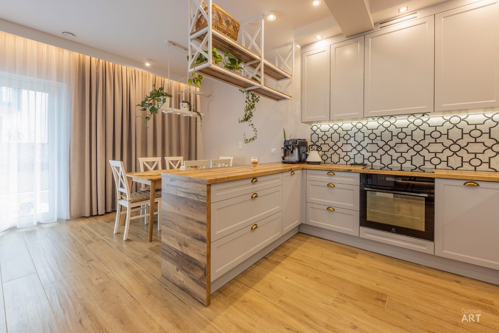 Nowoczesna kuchnia w jasnych kolorach z białymi szafkami, drewnianym blatem i wyspą kuchenną. Na ścianie widoczne są otwarte półki z roślinami, a w tle stół jadalny z krzesłami przy oknie zasłoniętym beżowymi zasłonami.