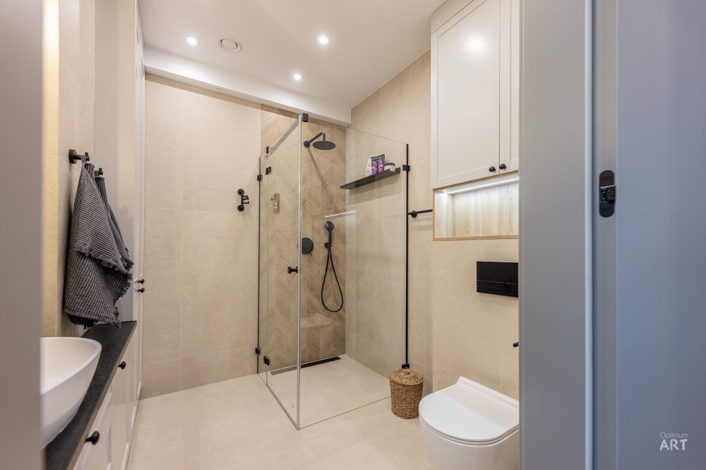 Nowoczesna łazienka z jasną kolorystyką, wyposażona w kabinę prysznicową ze szklanymi ściankami, białą miskę WC oraz umywalkę. Pomieszczenie ma białe meble łazienkowe i beżowe płytki na podłodze i ścianach.