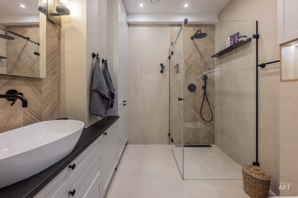 Nowoczesna łazienka z białą umywalką nablatową, czarną armaturą i szklaną kabiną prysznicową. Ściany wykończone są jasną płytką z wzorem jodełki, a na podłodze stoi wiklinowy koszyk.