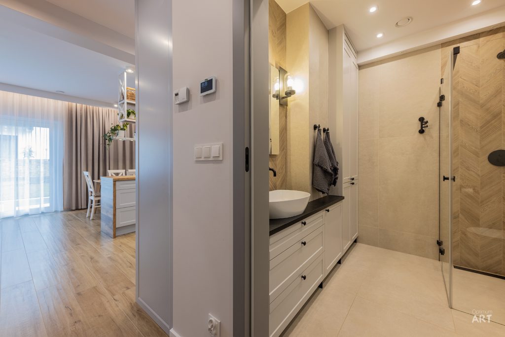 Nowoczesna łazienka z białymi szafkami, okrągłą umywalką na ciemnym blacie i szklaną kabiną prysznicową z drewnianym wzorem. W tle widoczny salon z drewnianą podłogą i oknem z zasłonami.