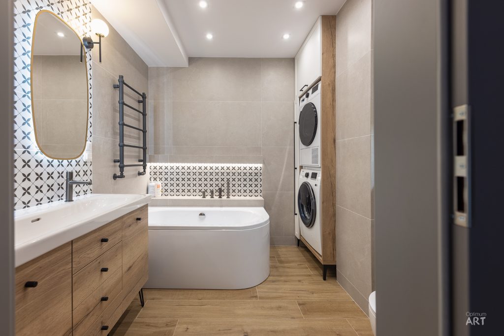 Nowoczesna łazienka z wanną wolnostojącą, drewnianą szafką z umywalką oraz pralką i suszarką w zabudowie. Ściany wykończone są beżowymi płytkami, a podłoga drewnopodobnymi płytkami, z dekoracyjnymi wzorami geometrycznymi na ścianie za lustrem.
