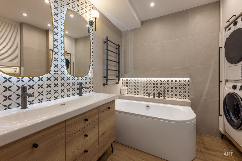 Nowoczesna łazienka z drewnianą szafką pod umywalkę, białą wanną narożną i geometrycznymi płytkami na ścianie. Po prawej stronie znajduje się pralka i suszarka, a nad wanną zamontowany jest czarny grzejnik.