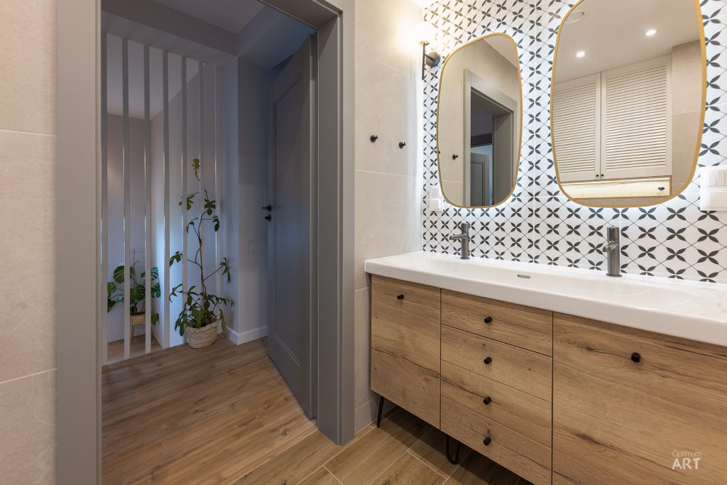 Nowoczesna łazienka z drewnianą szafką pod umywalkę, okrągłym lustrem w złotej ramie i charakterystyczną ścianą z białych płytek z czarnym wzorem geometrycznym. Po lewej stronie widoczne są szare drzwi prowadzące do innego pomieszczenia, a na podłodze znajduje się doniczka z rośliną.