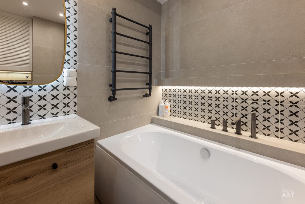 Nowoczesna łazienka z białą wanną, umywalką na drewnianej szafce i czarnym grzejnikiem ściennym. Ściany wykończone są beżowymi płytkami i białymi kafelkami z czarnym wzorem geometrycznym.