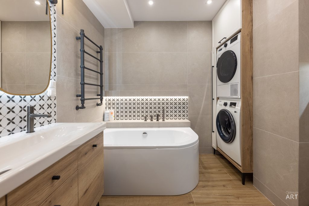 Nowoczesna łazienka z wanną wolnostojącą, drewnianą szafką z umywalką oraz białymi urządzeniami AGD (pralka i suszarka) umieszczonymi w zabudowie. Wnętrze wykończone jest beżowymi płytkami, drewnopodobnymi panelami na podłodze oraz dekoracyjnymi płytkami z geometrycznym wzorem.