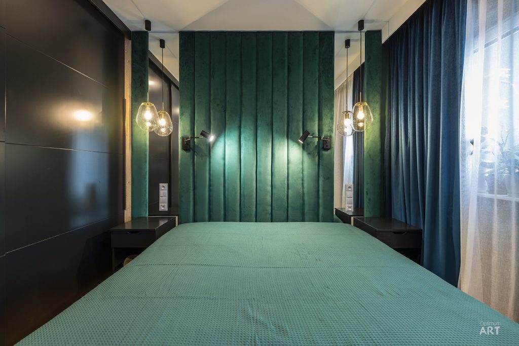 Nowoczesna sypialnia z łóżkiem pokrytym zielonym narzutem, na tle ciemnozielonej ściany z pionowymi panelami i czarnymi elementami po bokach. Pomieszczenie oświetlają szklane lampy wiszące oraz kinkiety, a po prawej stronie znajduje się okno z niebieskimi zasłonami.