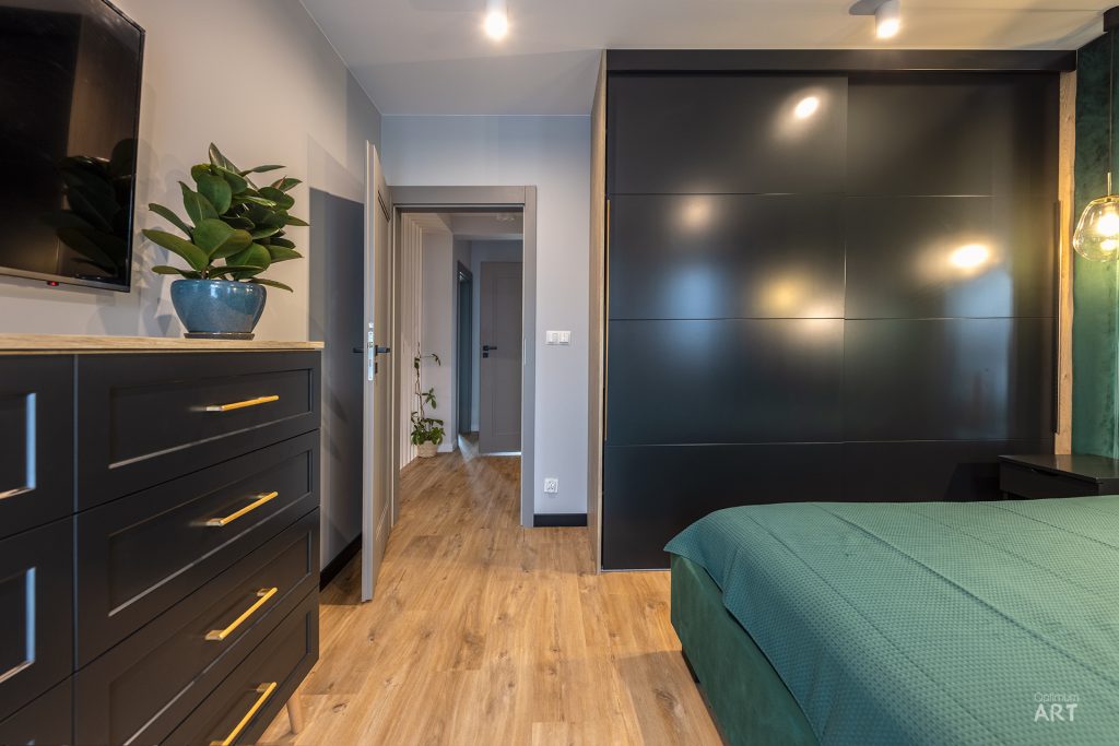 Nowoczesna sypialnia z ciemnymi meblami, łóżkiem z zielonym kocem i dużą czarną szafą. Na drewnianej podłodze widać komódkę z telewizorem i rośliną, a w tle drzwi prowadzące do korytarza.