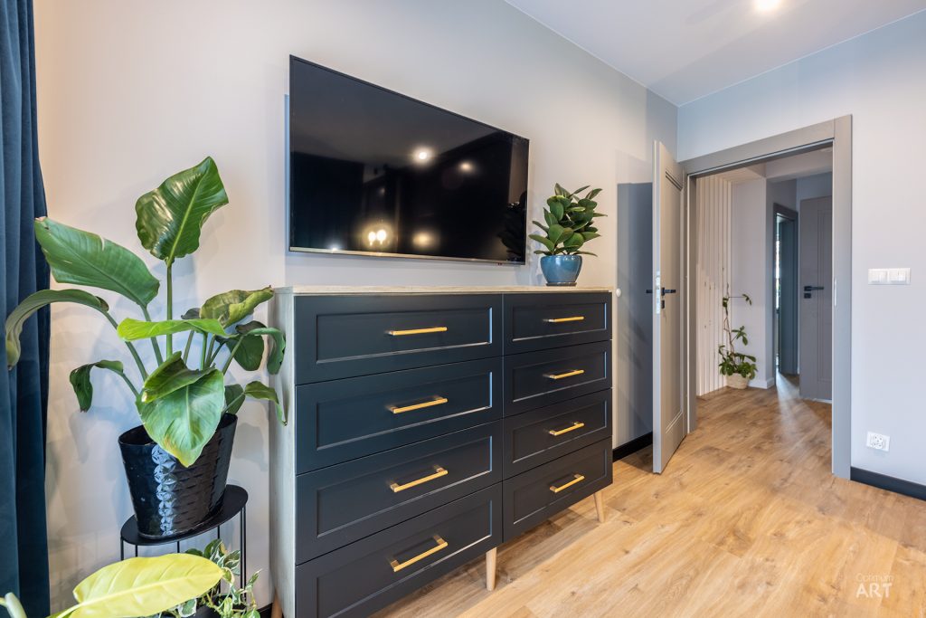 Nowoczesna sypialnia z ciemnoszarą komodą z wieloma szufladami, na której stoi duży telewizor. Po lewej stronie znajdują się donice z dużymi zielonymi roślinami, a w tle widać otwarte drzwi prowadzące do innych pomieszczeń.