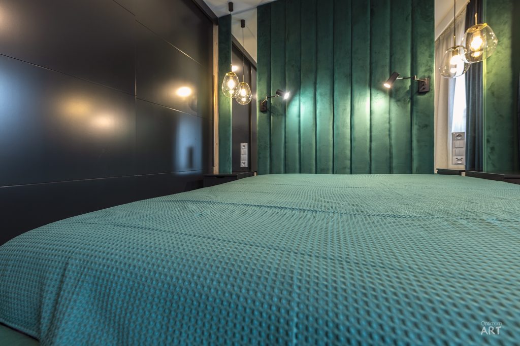 Nowoczesna sypialnia z łóżkiem przykrytym zieloną narzutą o teksturowanej powierzchni. Ściana za łóżkiem wykończona jest zielonymi panelami drewnianymi, a po bokach wiszą stylowe lampy wiszące ze szklanymi kloszami.