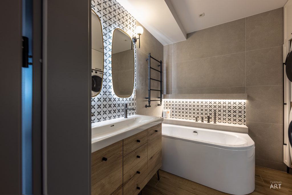 Nowoczesna łazienka z białą wanną wolnostojącą, drewnianą szafką pod umywalką i czarno-białymi płytkami w geometryczny wzór na ścianie. Pomieszczenie ma beżowe kafelki na pozostałych ścianach, lustro z podświetleniem i czarny grzejnik drabinkowy.