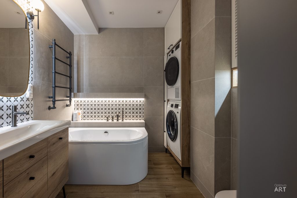 Nowoczesna łazienka z wanną wolnostojącą, drewnianą szafką pod umywalką i pralko-suszarką w zabudowie. Wnętrze utrzymane w beżowo-brązowej kolorystyce z dekoracyjnymi płytkami nad wanną.