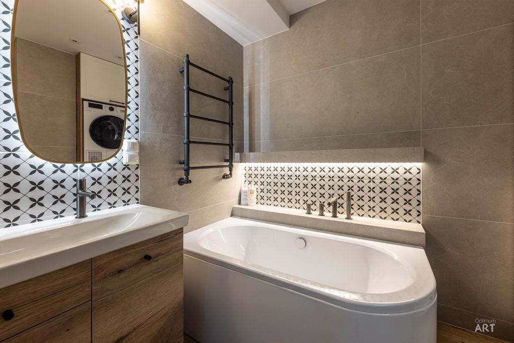 Nowoczesna łazienka z białą wanną narożną, drewnianą szafką pod umywalką i okrągłym lustrem. Na ścianie znajduje się czarny grzejnik drabinkowy, a w tle widać pralkę oraz ozdobne płytki z geometrycznym wzorem.