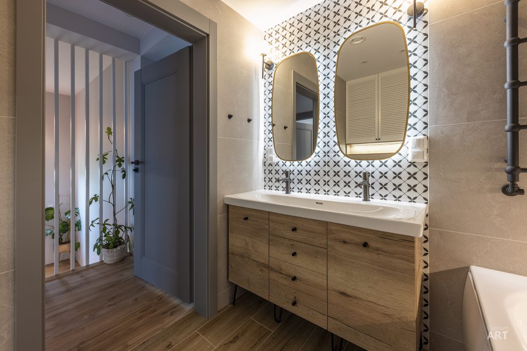 Nowoczesna łazienka z drewnianą szafką pod umywalką, okrągłym lustrem i charakterystyczną czarno-białą tapetą w geometryczny wzór na ścianie. Po lewej stronie widoczne są otwarte drzwi prowadzące do kolejnego pomieszczenia z drewnianą podłogą.