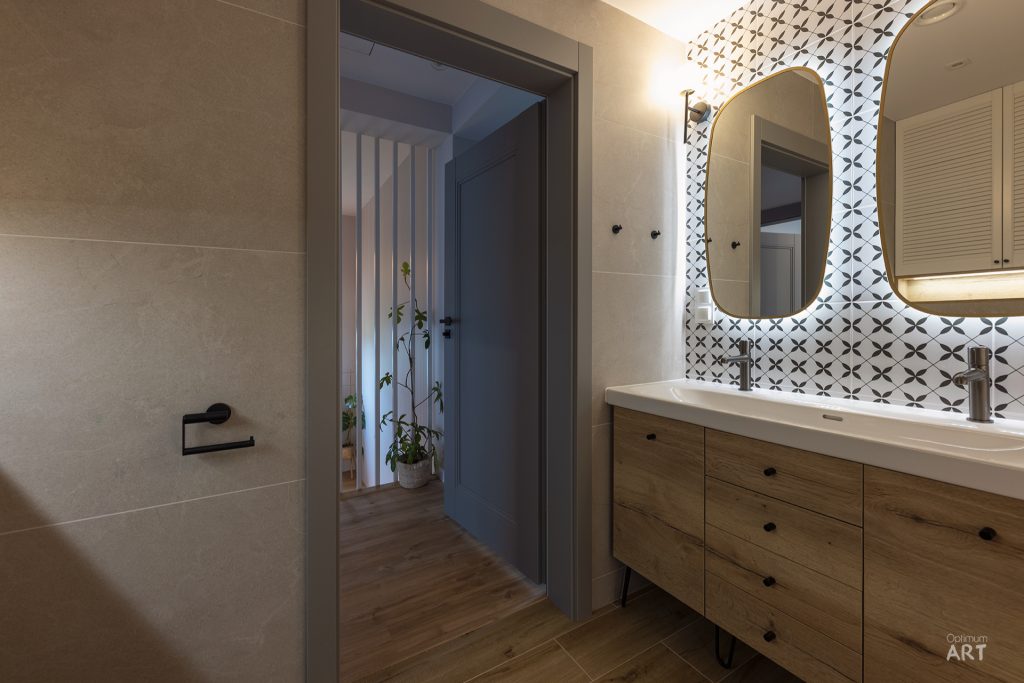 Nowoczesna łazienka z drewnianą szafką pod umywalką, okrągłym lustrem i dekoracyjnymi płytkami w geometryczny wzór na ścianie. W tle widać otwarte drzwi prowadzące do kolejnego pomieszczenia z drewnianą podłogą.