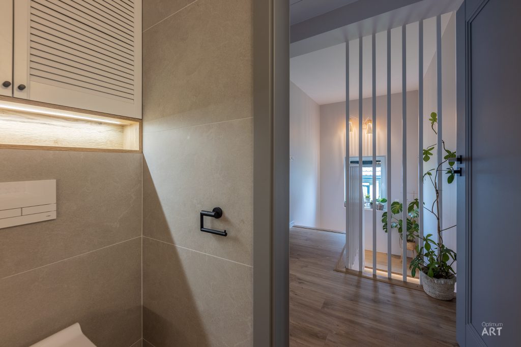 Nowoczesne wnętrze mieszkania z łazienką po lewej stronie wyłożoną beżowymi płytkami i salonem po prawej z drewnianą podłogą. Pomieszczenia dzieli stylowa przegroda z pionowych listew, przy której stoi roślina doniczkowa.