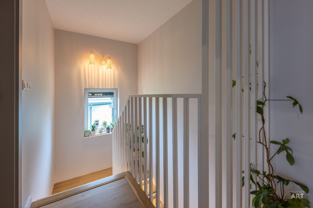 Wnętrze domu z drewnianymi schodami i białą balustradą z pionowymi szczeblami. W tle widać okno z roślinami na parapecie i wiszące lampy oświetlające przestrzeń.