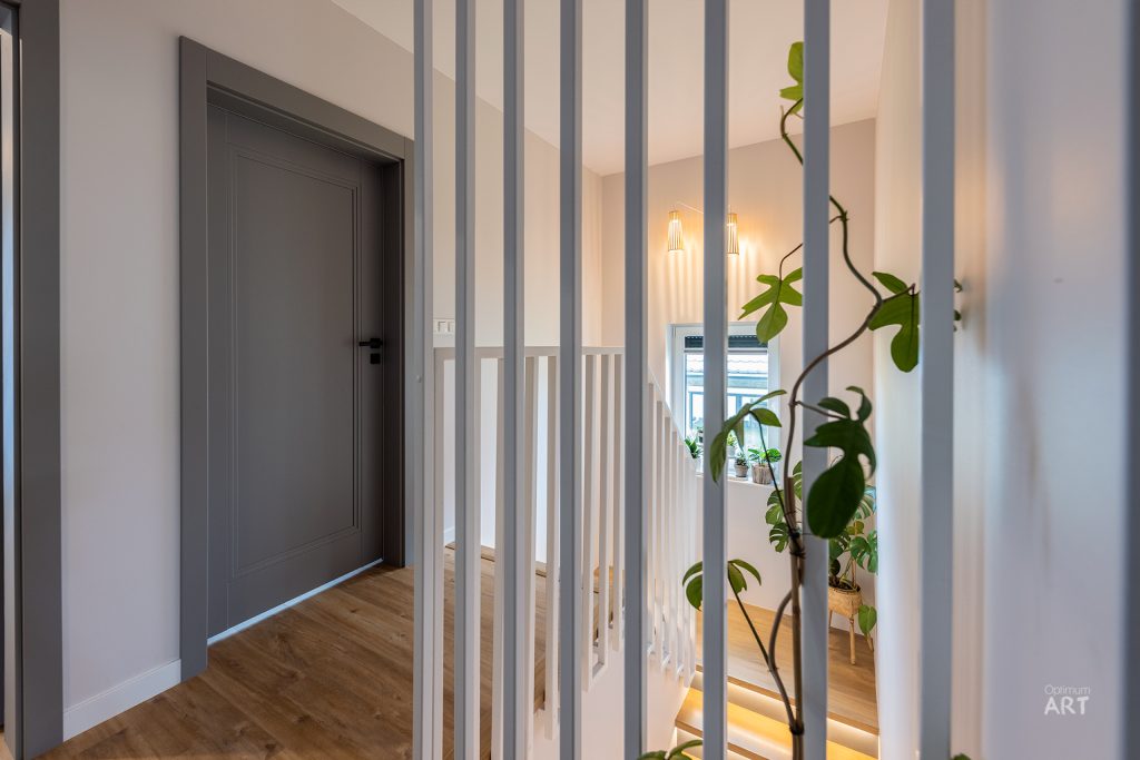 Nowoczesne wnętrze z szarymi drzwiami po lewej stronie i białą balustradą z pionowymi prętami po prawej. W tle widać rośliny doniczkowe i ciepłe oświetlenie, a podłoga wykonana jest z jasnego drewna.