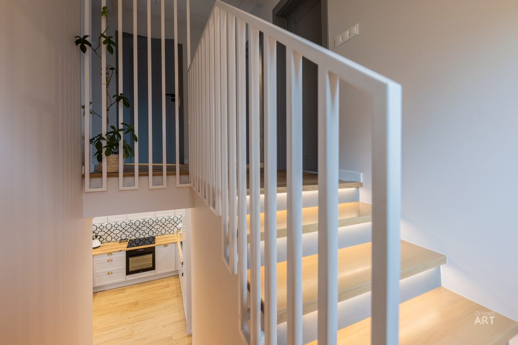 Białe drewniane schody z podświetlonymi stopniami prowadzą na górę, a przez balustrady widać kuchnię z jasnymi szafkami i wzorzystymi płytkami na ścianie. W tle znajduje się roślina doniczkowa i jasne wnętrze w nowoczesnym stylu.