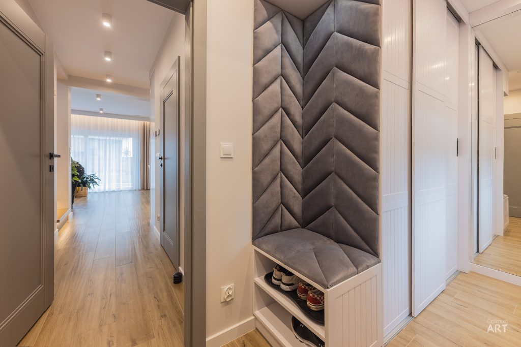 Nowoczesny przedpokój z jasnymi drewnianymi podłogami, białymi drzwiami i elegancką ławką z szarym pikowanym oparciem w kształcie jodełki. Na dole ławki znajduje się otwarta półka na buty, a przestrzeń jest dobrze oświetlona lampami sufitowymi.