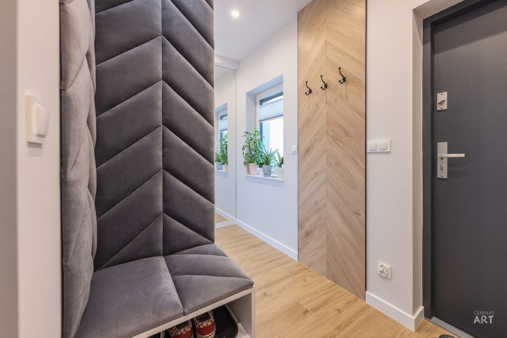 Nowoczesny korytarz z szarą tapicerowaną ławką przy ścianie, drewnianym panelem z wieszakami i ciemnymi drzwiami wejściowymi. W tle widać jasne pomieszczenie z roślinami doniczkowymi na parapecie okna.